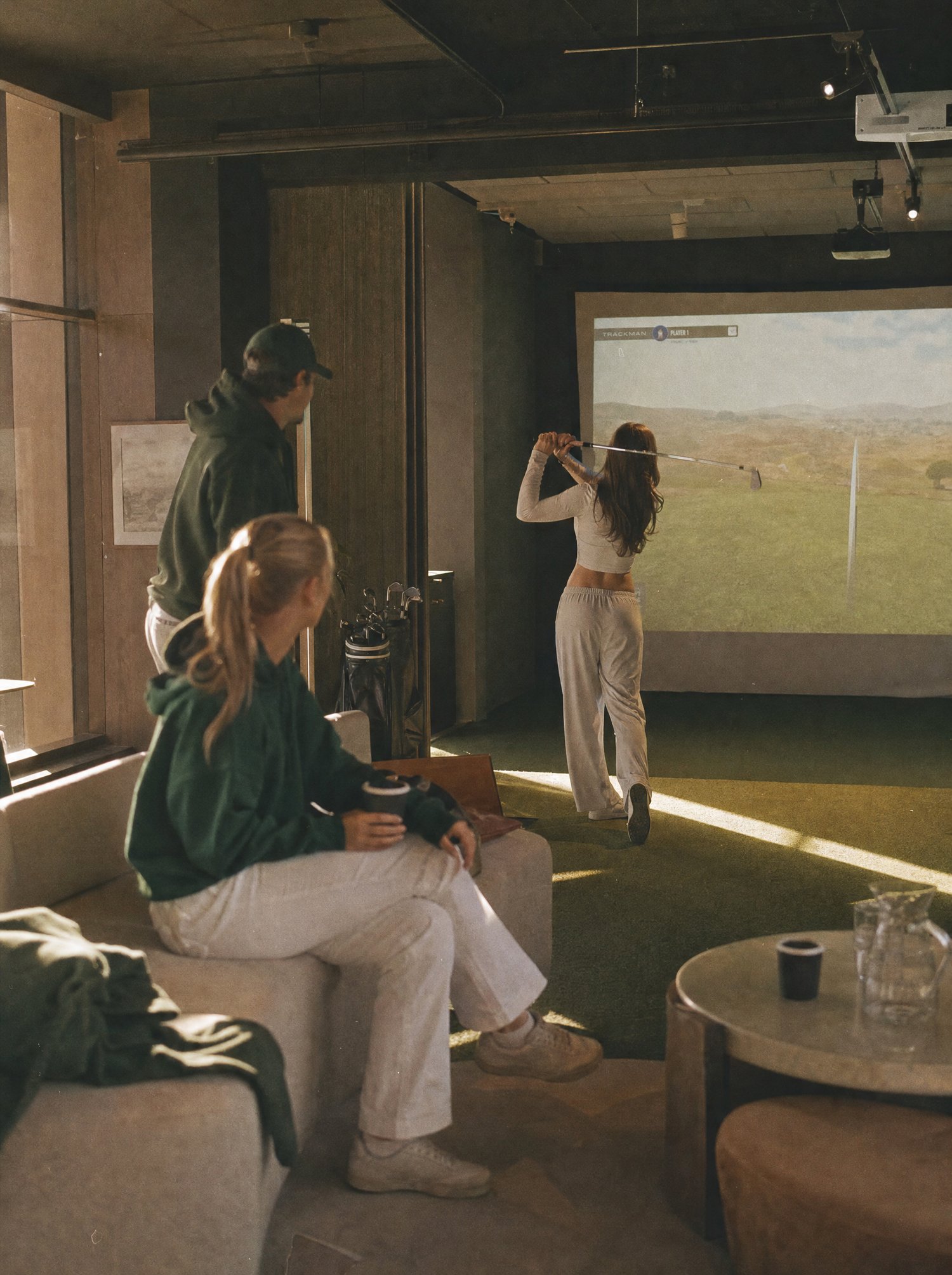 A golfer taking a swing on the simulator while two friends watch from the sofa